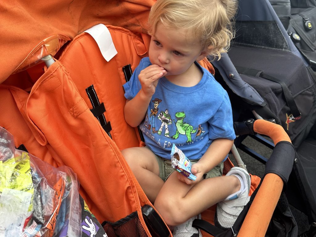 A young boy in a blue shirt eating a snack in an orange stroller. Taking snacks in, is another Disney World Tip