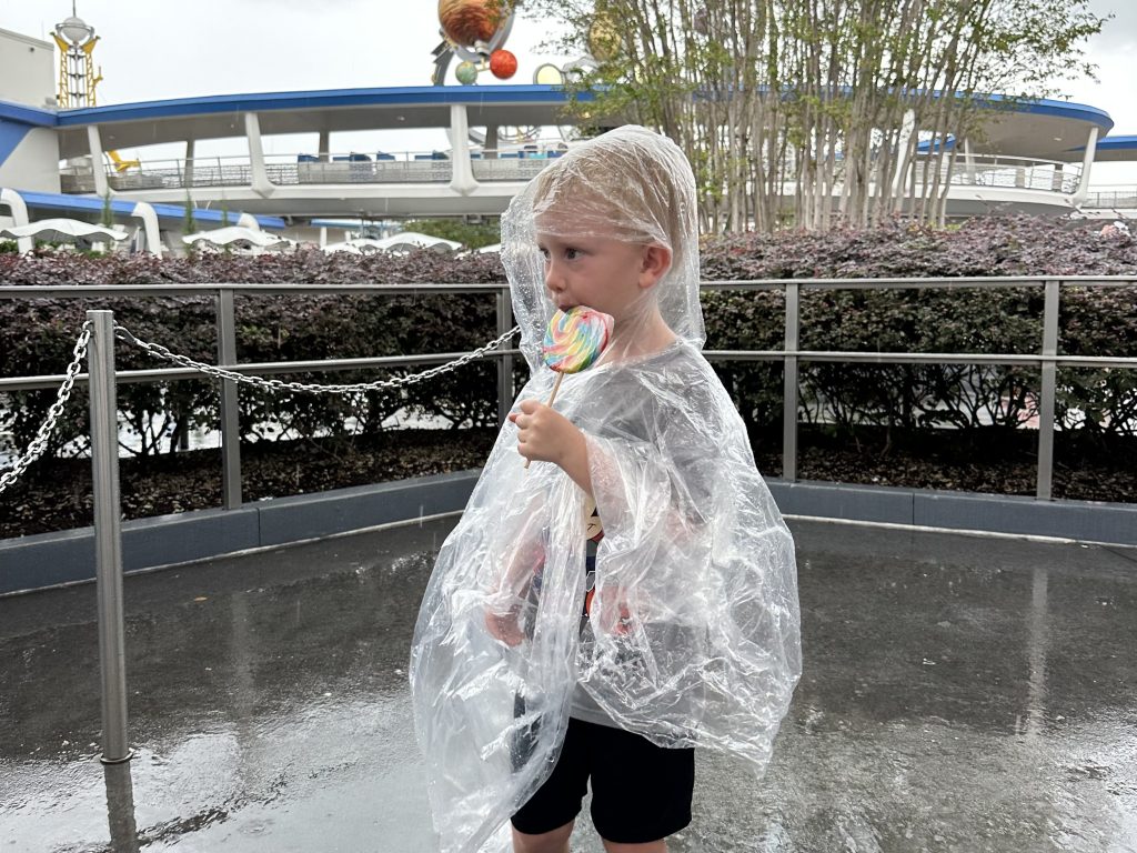 a young kid in a clear plastic poncho eating a giant lollipop at Disney World
