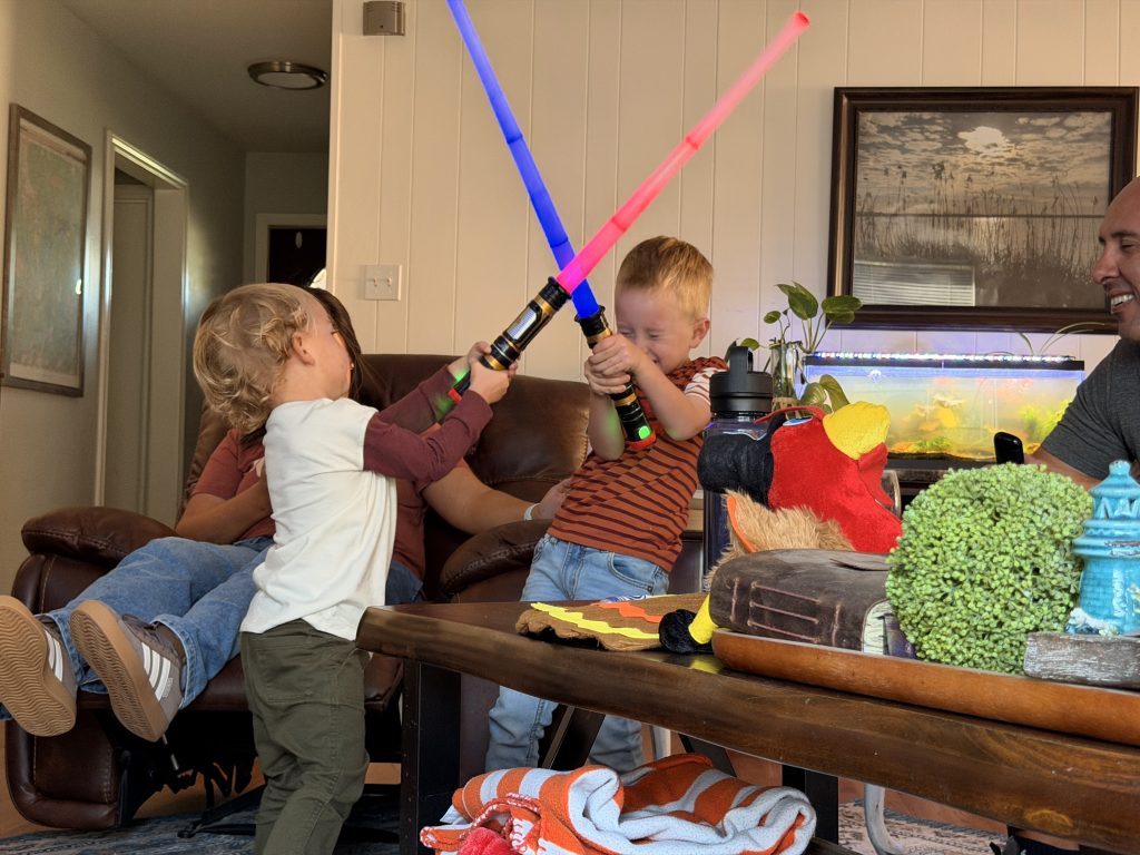 Two children playing with toy lightsabers in a living room. The child on the left is holding a blue lightsaber and wearing a white shirt and jeans. The child on the right is holding a pink lightsaber and wearing a striped shirt and jeans. A woman is sitting on a brown leather couch behind them, smiling. A man is sitting on the right side of the image, also smiling. A fish tank with colorful fish is in the background. A wooden table in the foreground has a toy car, a green plant, and a blue water bottle.