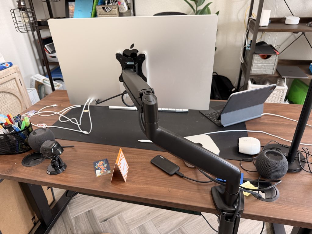 A wooden desk with a black monitor arm holding a white monitor and a laptop. A black microphone is attached to the monitor arm. A white mouse and a black smartphone are on the desk. A black speaker and a black circular object are also on the desk. A pen holder with pens and pencils is on the left side of the desk. A small card with a colorful design is on the desk. A white cable is on the desk. A black bag is on the floor behind the desk. A white wall is in the background.