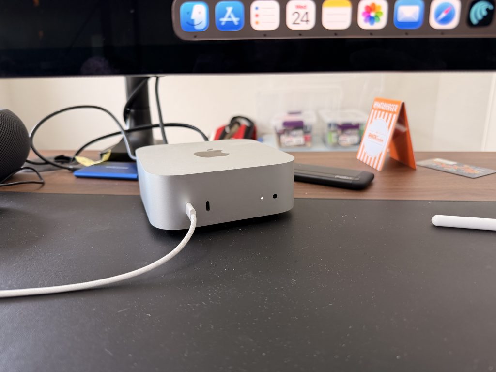A silver Apple M4 Mac mini on a black desk with a white cable.