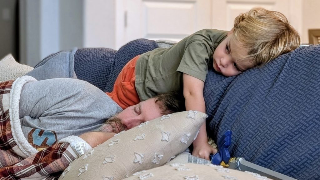 A man asleep with the flu on a blue couch. A small child is resting above him on the couch