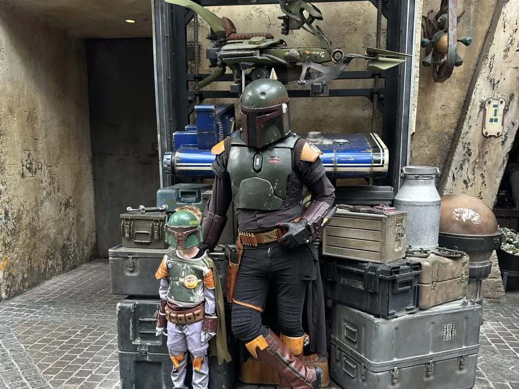 Two people in Boba Fett costumes standing next to crates and a spaceship model.