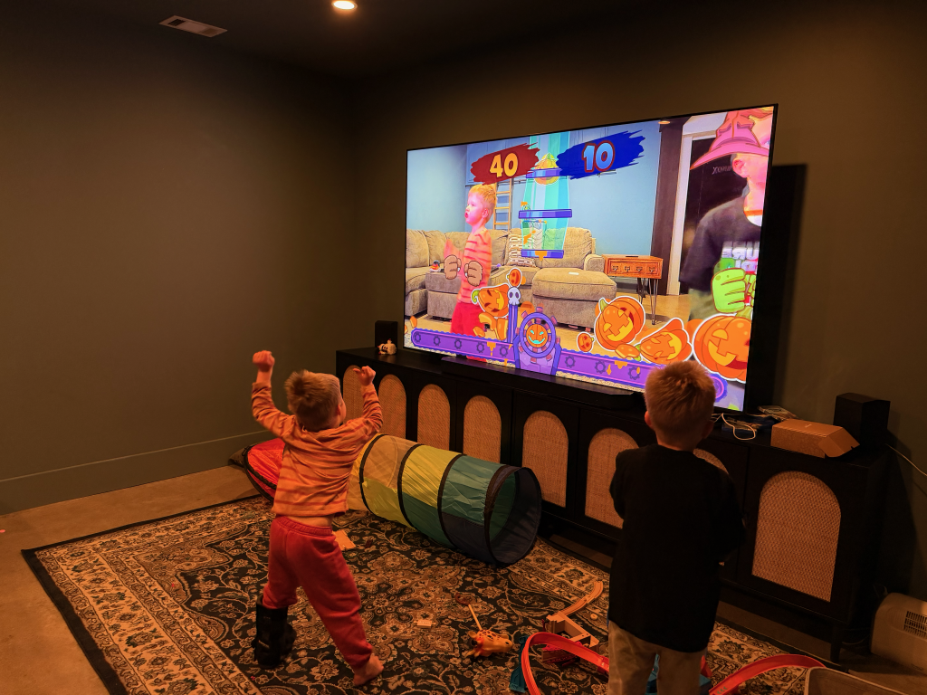 Two children playing a video game on a large screen in a room with toys and a rug.