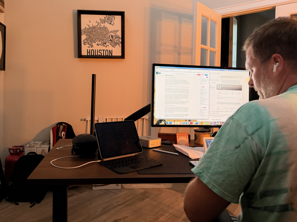 A man wearing earphones sits at a desk with a laptop and monitor, working in a room with a Houston map on the wall.