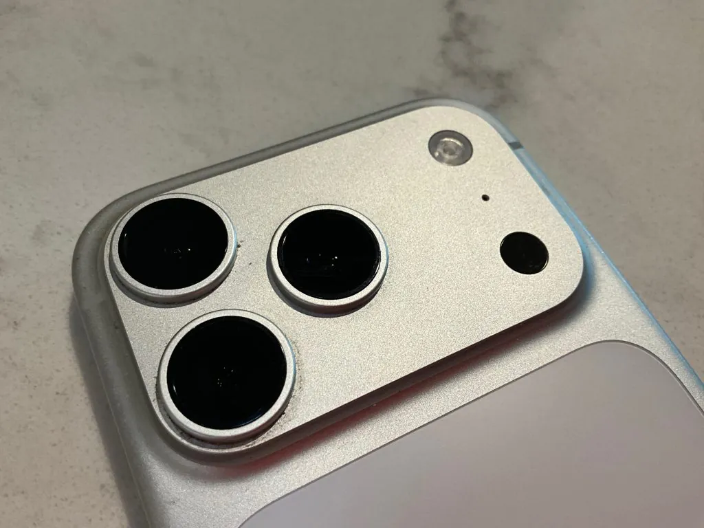 The iPhone 17 Pro One Year Later Review - An Amazing Device or No? 3 This image shows a close-up of the rear camera module on a silver iPhone 17. It features three distinct camera lenses arranged in a triangular pattern, along with a flash and a LiDAR sensor, all housed within a raised, square metallic bump.