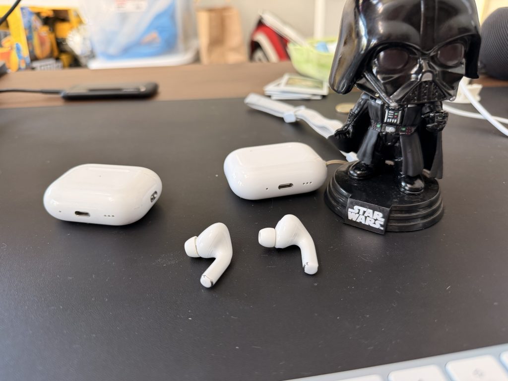 AirPods Pro 3 Review: Real-Life Impressions Of Fit, Sound Quality, Health Features, And More 4 AirPods Pro 2 case and an ear bud next to the AirPods Pro 3 case and an ear bud on a desk with a darth vader bobble head next to it.