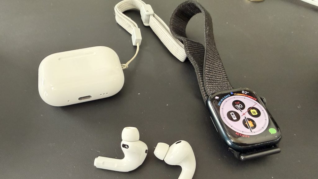 Why I Bought an Apple Watch Series 10 in 2025 (Even After Series 11 Launched) 4 An Apple Watch Series 8 with a black strap, a white AirPods Pro 3 charging case, and white earbuds on a gray surface.
