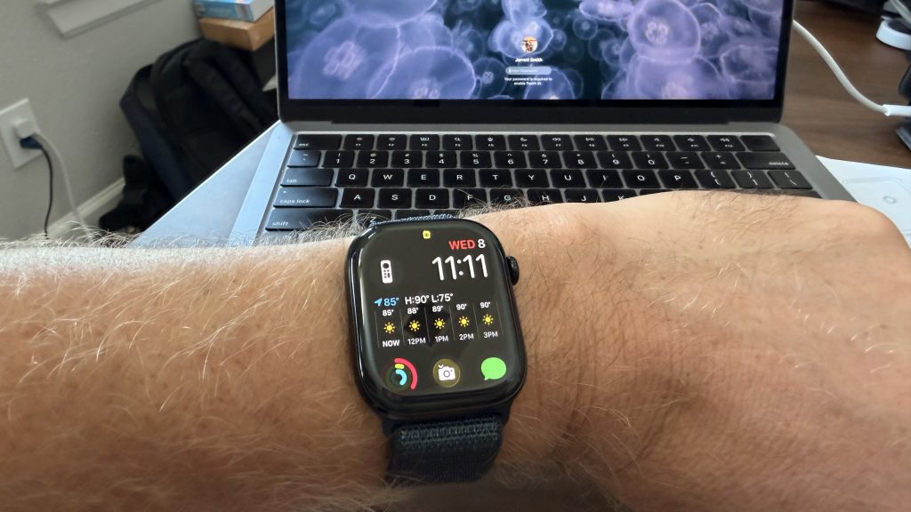 Why I Bought an Apple Watch Series 10 in 2025 (Even After Series 11 Launched) 3 A person wearing a black Apple Watch Series 10 on their left wrist. The watch face displays the time as 11:11, the date as Wednesday the 8th, and various app icons including weather, activity, and messages. The background shows a laptop with a colorful wallpaper and a black backpack on a wooden floor.