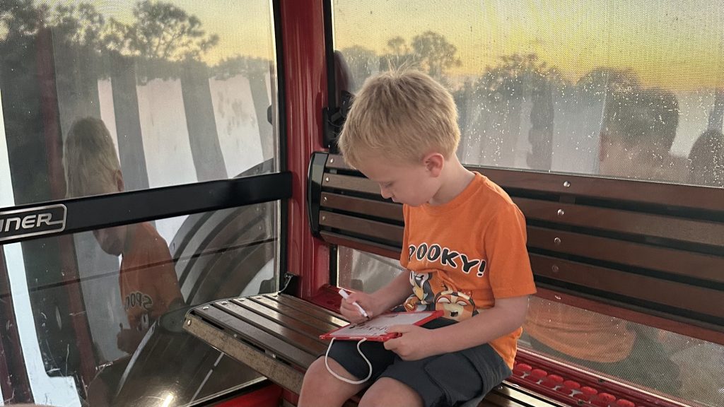 Conquering Disney World— My First Visit in Almost 40 Years 11 A boy in an orange shirt writing on a red rimmed handle-held white board sitting on the Skyliner at Disney World