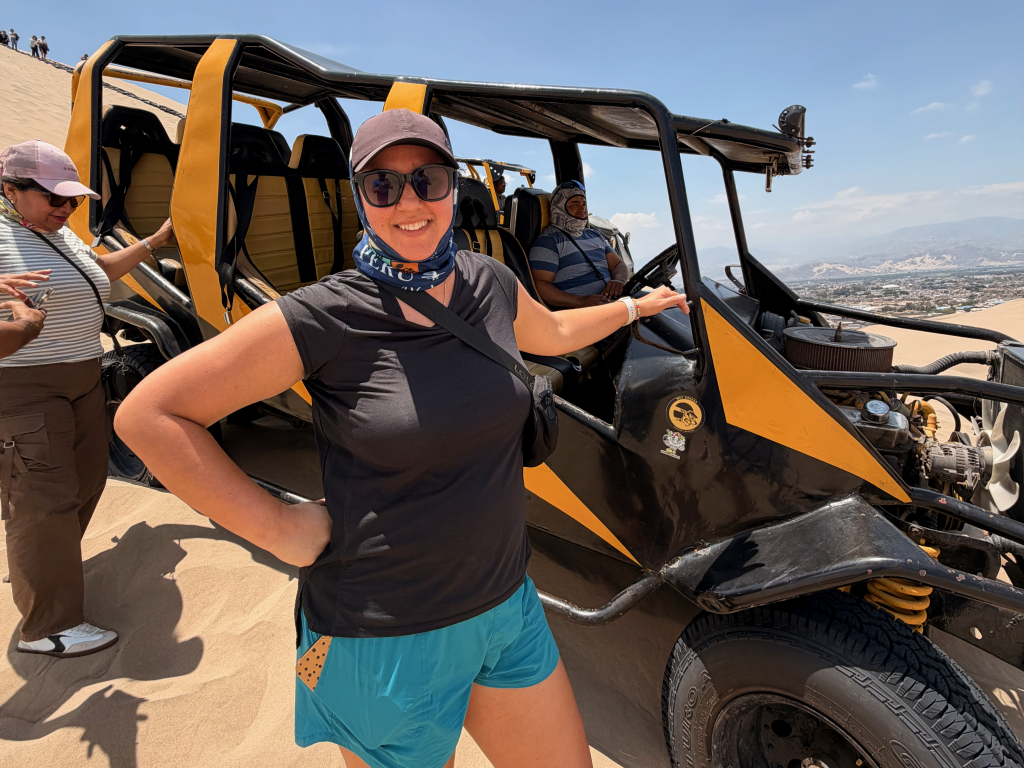 A woman in sunglasses and a cap poses next to a black and yellow dune buggy in a sandy desert.