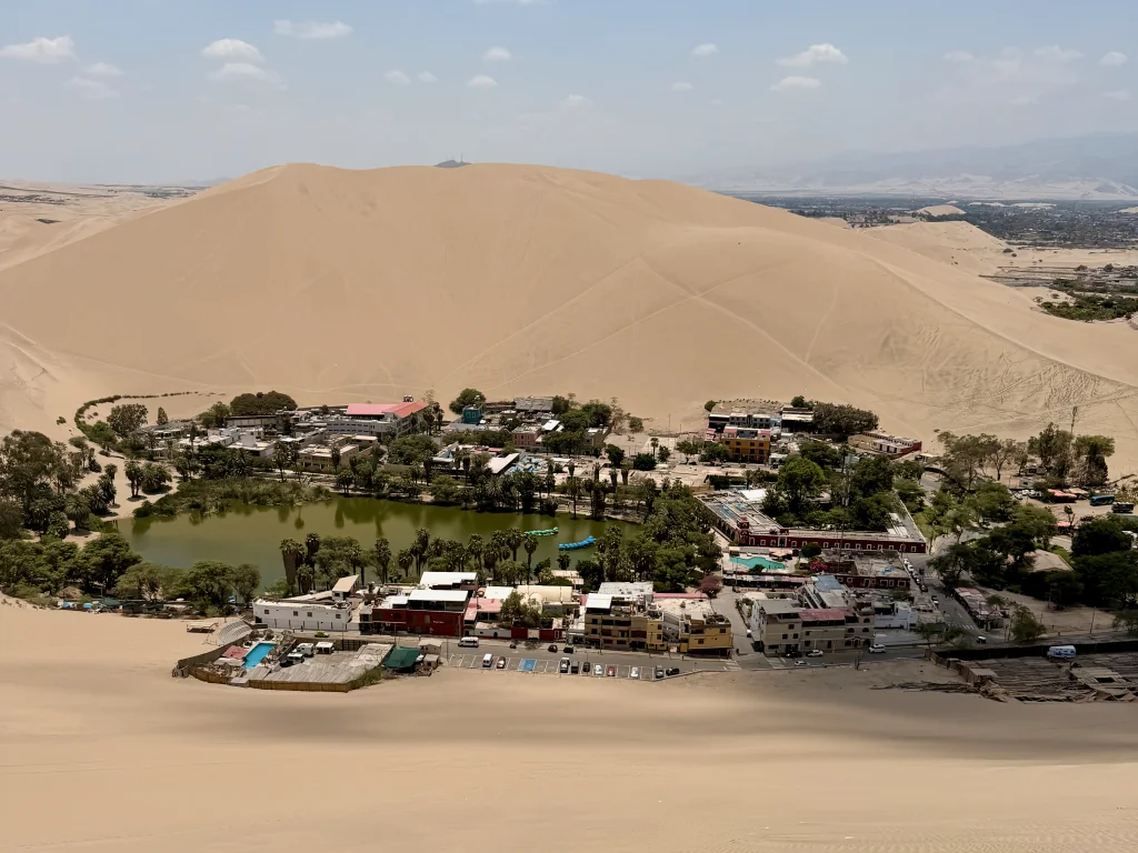 A green lake surrounded by buildings and sand dunes under a blue sky. The city of Huacachina, Peru