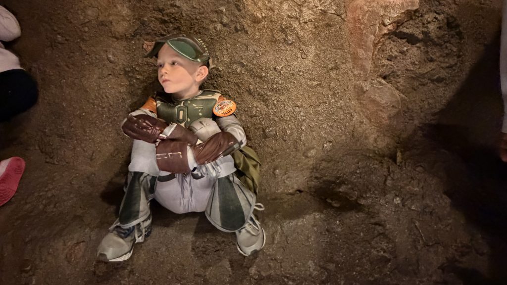 Child in Mandalorian costume sitting on rocky ground, looking contemplative.