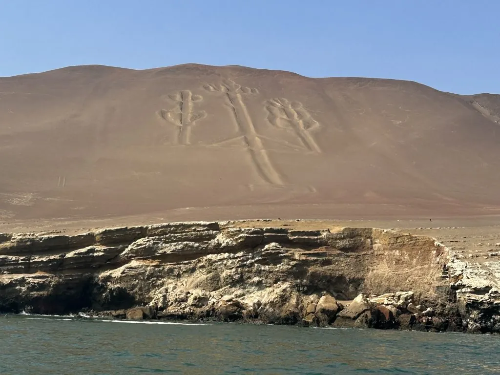 Geoglyphs of candelabra on a sandy hill beside a rocky shoreline under a clear blue sky.