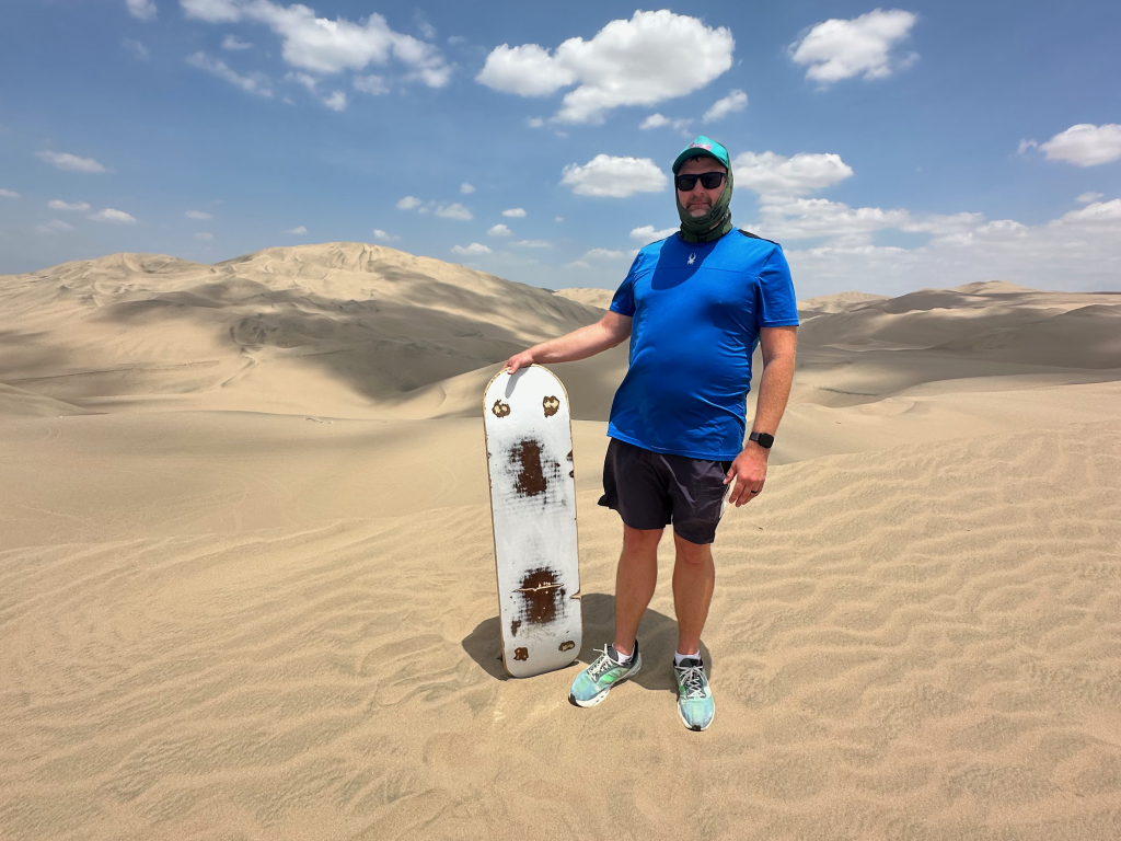 Man in blue shirt and sunglasses holding a sandboard in a desert.
