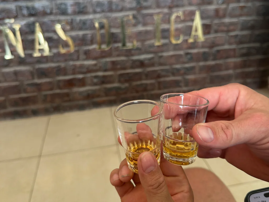 Two people holding shot glasses of amber liquid in front of a brick wall with gold lettering.