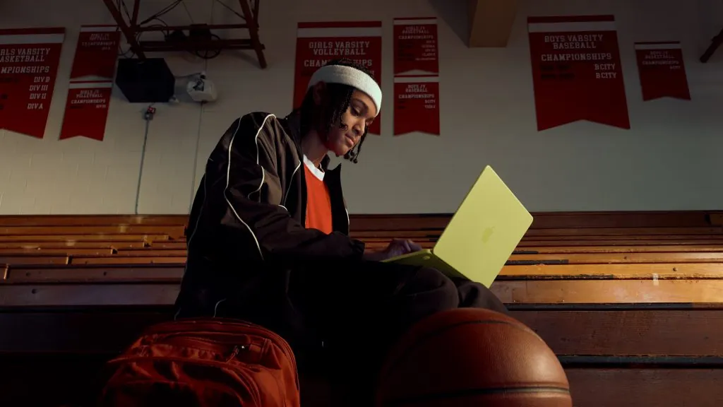 A person in a white cap sitting on bleachers, using a citrus MacBook Neo, with basketballs nearby.
