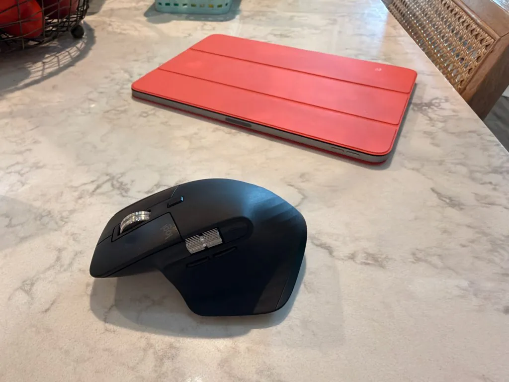 This image features a black Logitech MX Master 3 mouse resting on a light-colored, marble-patterned surface. In the background, there is a closed tablet inside a bright red case
