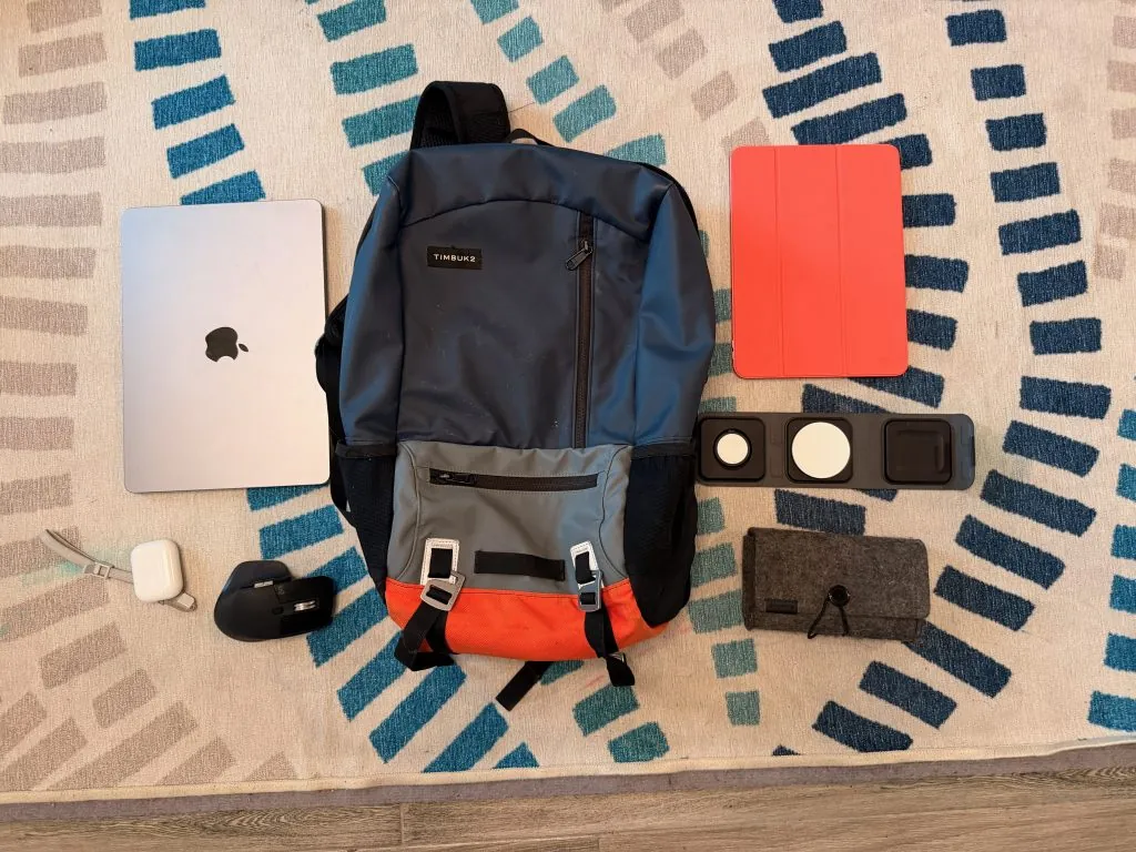 A writer's travel bag, specifically a Timbuk2 backpack, surrounded by a MacBook, an iPad with a red cover, AirPods, a Logitech mouse, a 3-in-1 charging pad, and a gray felt pouch, all laid out on a patterned rug.
