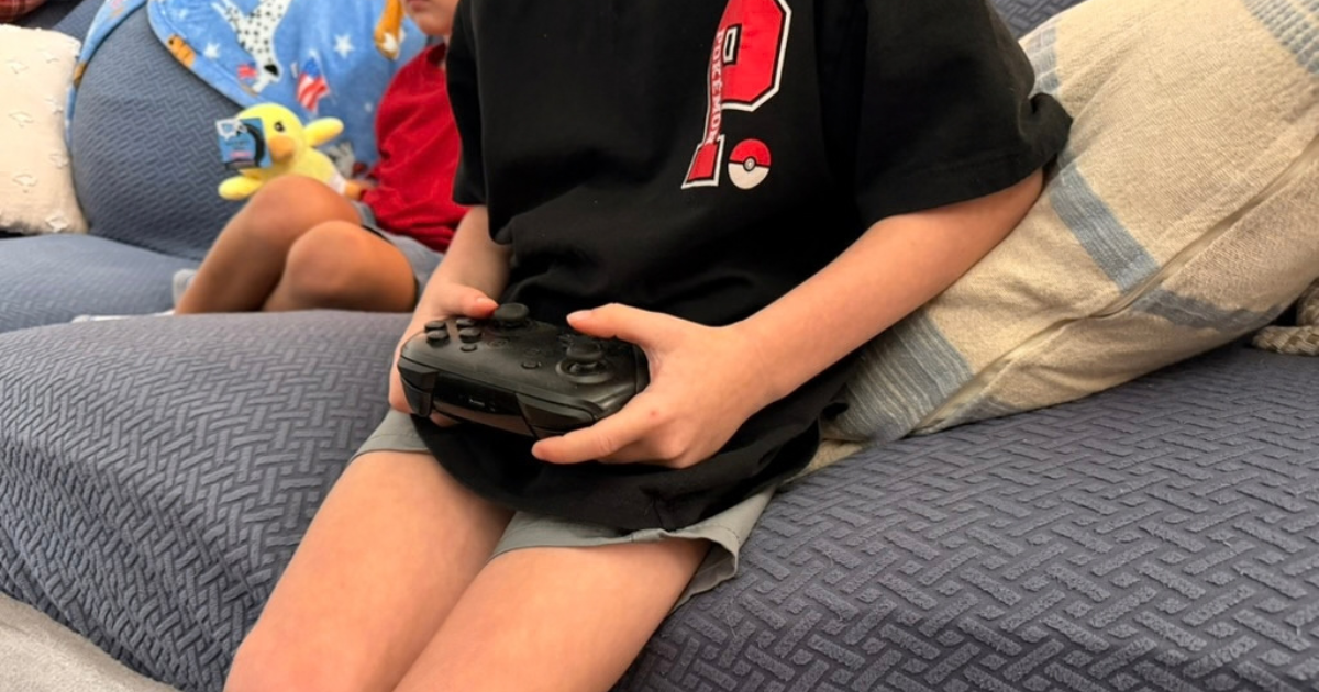 A close-up of a child sitting on a blue couch, holding a black video game controller. This image highlights the connection between video games and kids, showing a casual moment of play at home.
