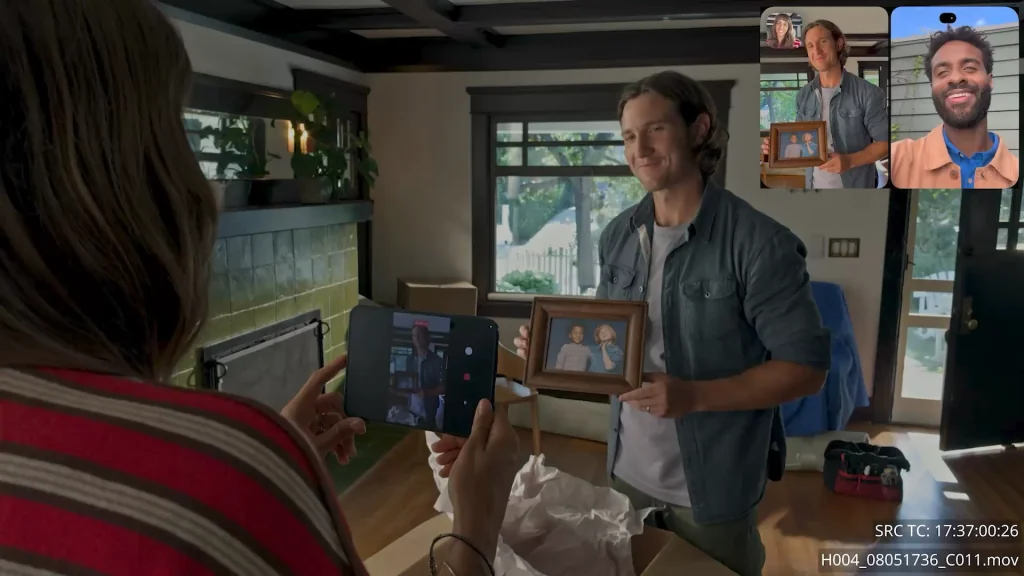 A person holding an iPhone, taking a photo of a framed picture held by a smiling man, with another man visible on the phone's screen in a video call.
